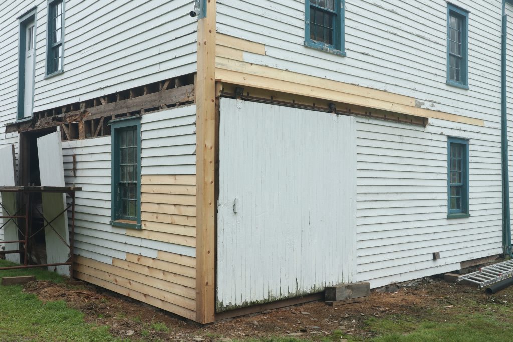 The exterior corner of a white building being repaired.  Several siding pieces and the corner piece have been replaced.  More siding pieces will be replaced and painted white.