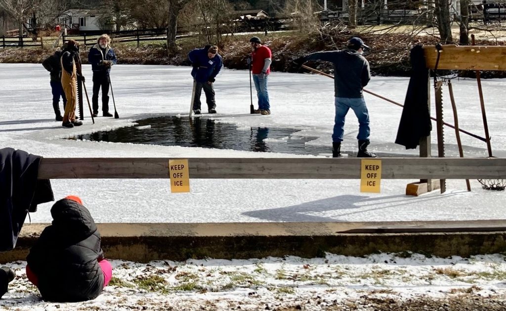 Men working around a hole in the frozen surface of a pond cutting blocks of ice and pushing them on shore.  A child watching in the foreground from behind a “keep off ice” sign.