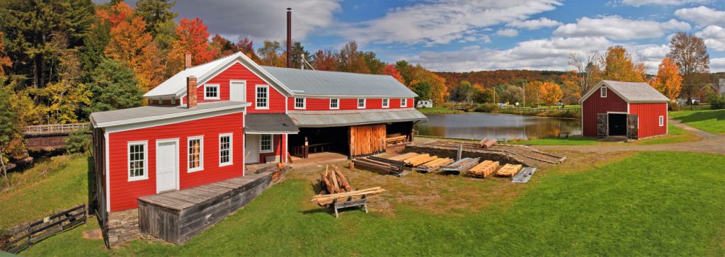 planks of wook in front of a red sawmill in the foreground with a red shed to the right and a pond in the background