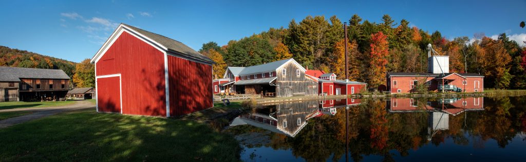 a red shed in the left forground sits on the bank of a pond with natural wood and red buildings and trees in the background
