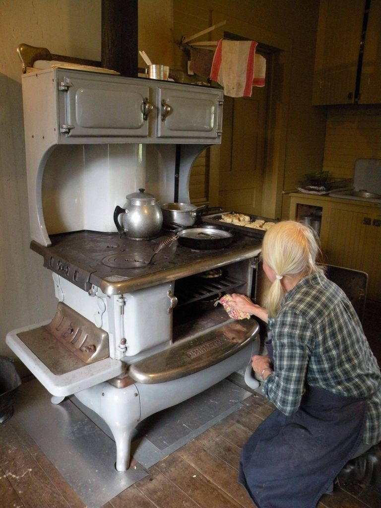 a woman kneels in front of a wood-fire cookstove and reaches into the oven